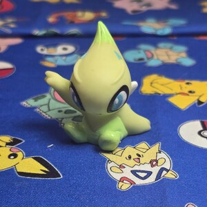 Puede incluir: Una figura Pokémon verde claro y amarilla con ojos azules y una cabeza puntiaguda. La figura está sentada sobre una tela azul estampada con varios personajes de Pokémon. La figura tiene una expresión amigable y los brazos extendidos.
