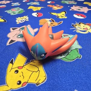 以下が含まれることがあります： オレンジと青色の星型のポケモンフィギュア。様々なポケモンキャラクターのデザインが施された青色の生地の上に置かれています。フィギュアは濃いグレーのアクセントと青い顔をしています。生地にはピカチュウやフシギダネなどのキャラクターが描かれています。