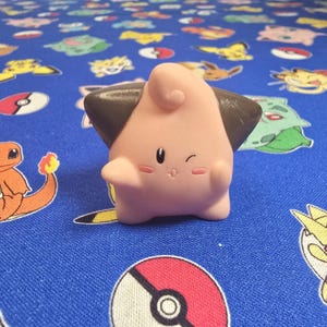 May include: A pink Clefairy figurine with brown ear tips and a small, smiling face. The figurine is on a blue fabric patterned with various cartoon characters.