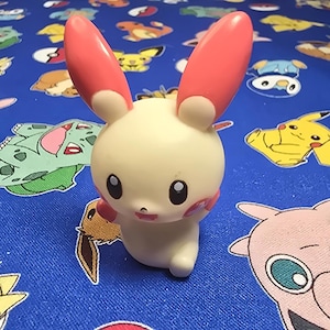 May include: A cream-colored, bunny-like figurine with large, pink-tipped ears and rosy cheeks. The figure has large black eyes and a small, smiling mouth. It is set against a blue background with various cartoon character designs.
