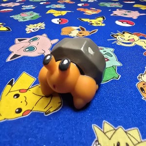 May include: An orange and gray toy hermit crab figurine with large black eyes. The figurine is on a blue fabric covered with various cartoon character designs.