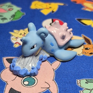 Puede incluir: Figura de Lapras azul claro con caparazón rosa y base blanca y azul, sobre tela azul estampada con personajes de dibujos animados. Es un artículo de colección.
