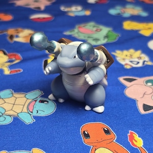 May include: A blue and gray Blastoise figurine with two blue cannons extending from its shell. The toy is on a blue fabric with various cartoon character designs.