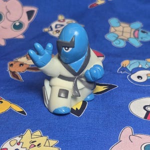 May include: A blue and white figurine of a character in a karate gi. The figure has a blue head and hands, a white gi with a gray belt, and is posed in a karate stance. The background is a blue fabric with cartoon characters.