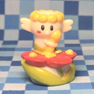 May include: A small figurine of a pale, winged creature with yellow hair, sitting inside a large red flower with green leaves. The creature has large black eyes, a small red cheek mark, and is holding a yellow object against its chest.