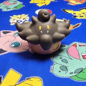 May include: A brown and pink Pokemon figure with yellow accents. The figure has a bulbous top and a rounded base with yellow spots. The background is a blue fabric with various Pokemon character designs.