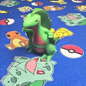 May include: A green and red Sceptile figurine stands on a blue surface decorated with various Pokémon characters and Poké Ball designs. The figurine has a detailed design, with a green body, red chest, and yellow eyes. The background features other Pokémon.