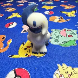 May include: A white and blue Absol figurine with red eyes, standing on a blue fabric patterned with various cartoon characters and Pokeballs. The Absol figure has a dark blue horn and face.