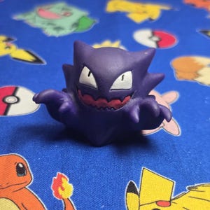 Può includere: Una statuetta viola di Haunter con occhi bianchi e bocca rossa. La statuetta è su una superficie blu a fantasia con altri personaggi dei cartoni animati.
