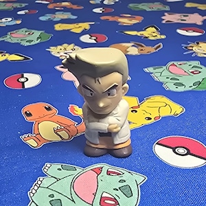 Puede incluir: Una pequeña figura de un personaje con cabello castaño claro y una bata blanca, de pie sobre una superficie azul. La superficie está cubierta con diseños de personajes de dibujos animados coloridos, incluido un Pokeball rojo y blanco.