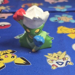 May include: A green and white figurine of a character with red and blue accents. The figurine is on a blue patterned surface with cartoon characters.