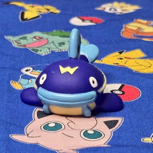 Könnte beinhalten: Eine blau-gelbe Pokémon-Figur mit einem gelben 'W' auf dem Kopf. Die Figur befindet sich auf einem blauen Hintergrund mit verschiedenen Pokémon-Charakterdesigns.