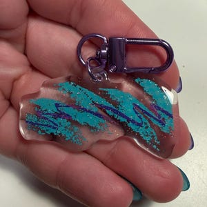 Może przedstawiać: Przezroczysty akrylowy brelok z fioletowym zapięciem. Brelok ma turkusowy i fioletowy abstrakcyjny wzór przypominający kubek jednorazowy. Brelok jest trzymany w dłoni.