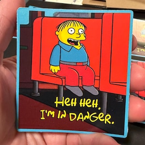 May include: A square sticker depicting a cartoon character with yellow skin, a blue shirt, and red trousers, seated in a red chair. The text "Heh heh, I'm in danger" is written in yellow. The sticker has a blue border.