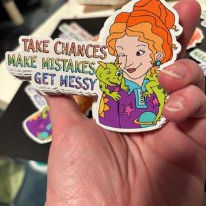 Pode incluir: Um autocolante colorido com uma mulher de desenho animado com cabelo laranja e um dragão verde. O autocolante tem o texto "TAKE CHANCES MAKE MISTAKES GET MESSY" em cores do arco-íris. A mulher está a usar uma camisa roxa com estrelas.