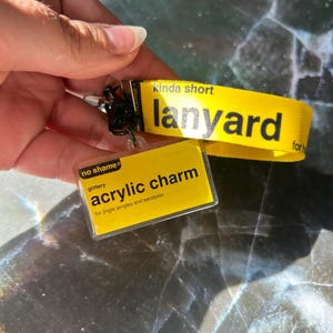 May include: A yellow lanyard with black text reading "lanyard". A yellow acrylic charm is attached, with the words "acrylic charm" and "glittery" in black. The charm reads "for jingle jangles and serotonin."