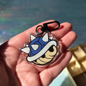 May include: A clear acrylic keychain with a cartoon character design. The character has a blue shell, white spikes, and a tan face. It includes a black clasp and ring for attachment. The design is outlined in black.