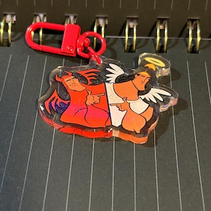 May include: A clear acrylic keychain featuring a cartoon devil and angel design. The devil is red with horns and a tail, while the angel has white wings and a halo. The keychain has a red clasp.