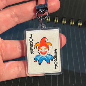 Puede incluir: Un llavero acrílico transparente con un diseño de carta de Joker pixelado. La tarjeta tiene un bufón sonriente con un sombrero rojo y un atuendo azul. La palabra "JOKER" está impresa verticalmente en ambos lados. El llavero tiene un cierre plateado.
