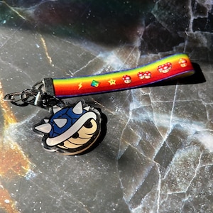 May include: A keychain with a blue spiky shell charm and a rainbow lanyard decorated with mushroom and star graphics. The lanyard is attached to a silver chain and clasp. The background is a dark, marbled surface.