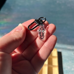 May include: A clear resin keychain charm shaped like a paperclip with a cartoon face. The charm has black eyes and a black mouth. It is attached to a black metal clasp.