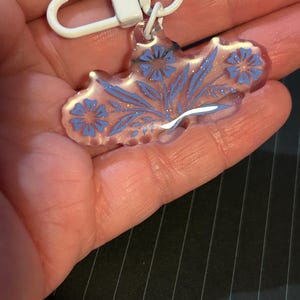 May include: A clear, floral-shaped keyring featuring blue flower and leaf designs. It has a white metal clasp and ring for attaching to keys or bags. The design includes glitter accents.