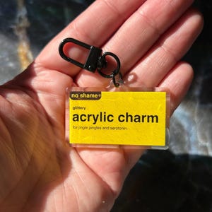 Könnte beinhalten: Ein klarer Acryl-Schlüsselanhänger mit gelbem Hintergrund und schwarzem Text, der "no shame" und "acrylic charm" lautet. Der Anhänger hat einen schwarzen Verschluss. Der Text unter den Worten "acrylic charm" lautet "for jingle jangles and serotonin".