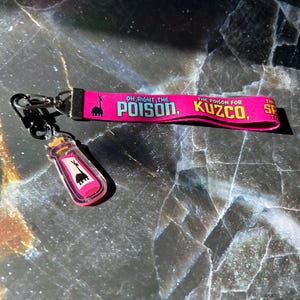 Puede incluir: Un juego de llavero y cordón rosa con el texto "OH, RIGHT, THE POISON, KUZCO" en blanco y negro. El llavero presenta un pequeño vial transparente con líquido rosa y un gráfico de llama.