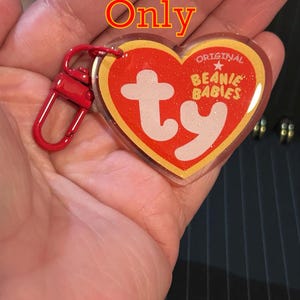 May include: A red heart-shaped keychain with the text "ty" in white and the words "Original Beanie Babies". The keychain has a red clasp and the text "Keychain Only" in the upper part of the image.