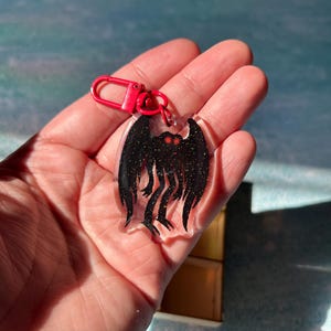 Peut inclure: Un porte-clés en acrylique transparent avec un motif de créature noir rempli de paillettes et un fermoir rouge. Le motif présente des yeux rouges. Le porte-clés a un aspect unique et stylisé.