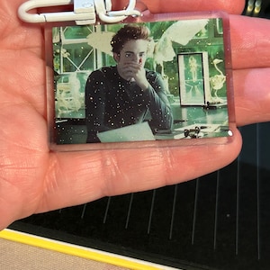 Puede incluir: Un llavero rectangular transparente con una foto de una persona con cabello oscuro, vistiendo una camisa oscura de manga larga. El llavero tiene un cierre de metal blanco. El fondo es una habitación de tonos verdes con obras de arte y un escritorio.