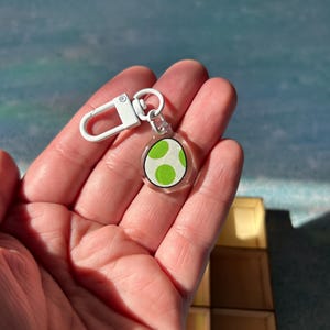 May include: A clear acrylic keyring with a green and white egg design, attached to a white metal clasp. The keyring is held in a hand against a blurred background.