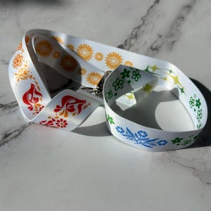 Puede incluir: Cordón blanco con coloridos estampados florales en rojo, naranja, amarillo, verde y azul. El cordón tiene un cierre de metal y está hecho de un material liso y satinado. Los diseños florales incluyen girasoles y otras flores estilizadas.