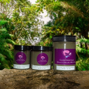 May include: Three glass jars of facial mask with black lids and labels. The labels are purple with white text that reads "Refreshing Peppermint Facial Mask" and the size in ounces. The jars are on a brown wooden surface.