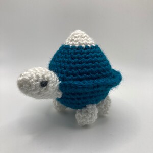 以下が含まれることがあります： 白い体とターコイズブルーの殻を持つ、かぎ針編みのかめのおもちゃ。殻の上には白い頂上があります。