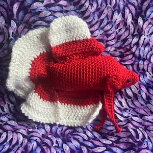 May include: A crocheted betta fish toy made with red and white yarn. The fish has a long tail and fins, and is posed on a purple background with a fish pattern.