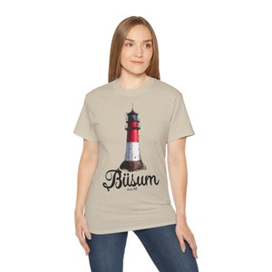 May include: A beige t-shirt with a graphic of a red and white lighthouse with a black roof and the text "Büsum" in black cursive lettering.