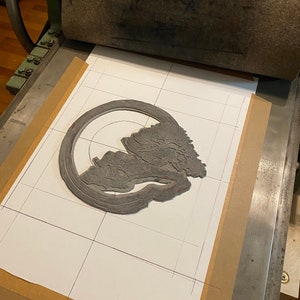 May include: A grey linoleum block print of a tree with a circular design around it. The print is on a white sheet of paper with a grid pattern drawn on it. The print is on a printing press.