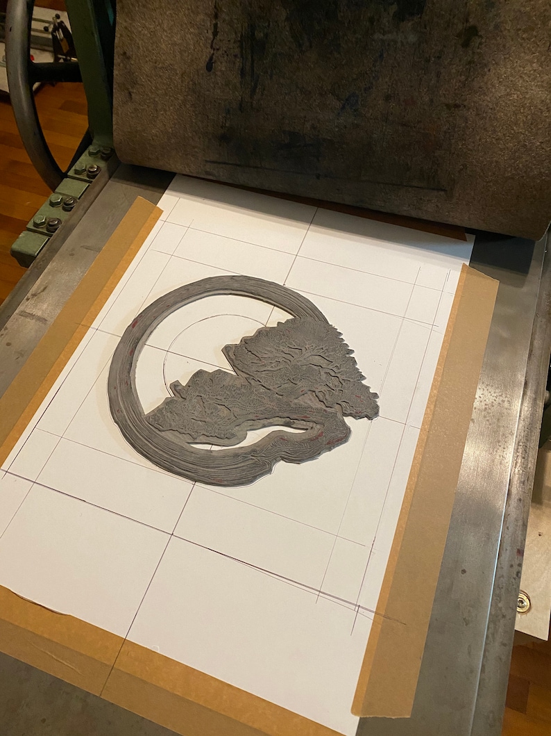 May include: A grey linoleum block print of a tree with a circular design around it. The print is on a white sheet of paper with a grid pattern drawn on it. The print is on a printing press.