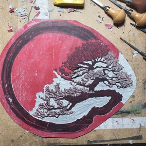May include: A red and white woodblock print of a pine tree with a black circle around it. The print is on a wooden surface with carving tools scattered around it.