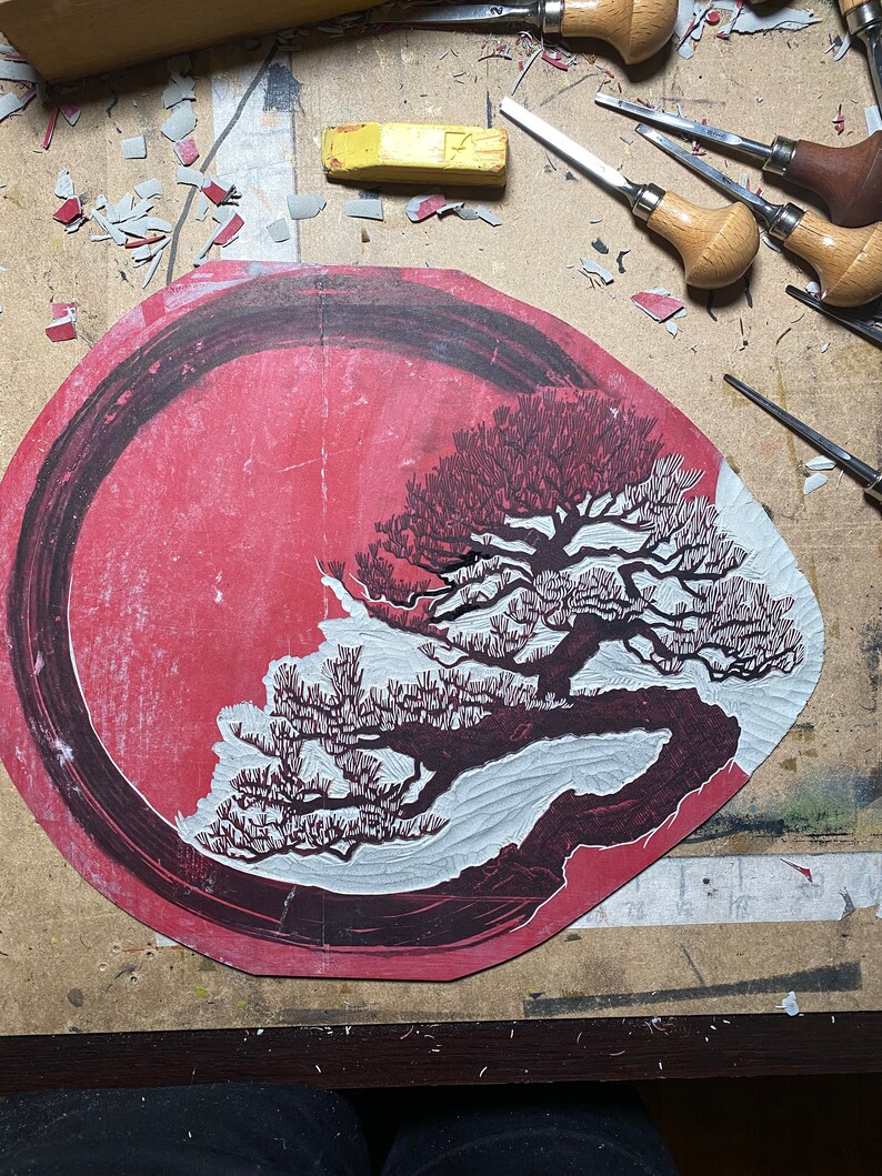 May include: A red and white woodblock print of a pine tree with a black circle around it. The print is on a wooden surface with carving tools scattered around it.