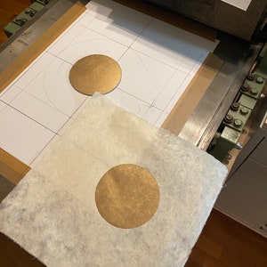 May include: A gold circle is being pressed onto a piece of white paper with a textured surface. The paper is on a printing press.