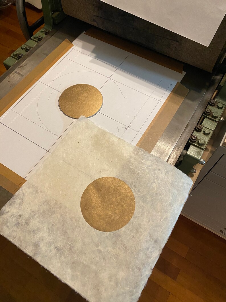 May include: A gold circle is being pressed onto a piece of white paper with a textured surface. The paper is on a printing press.