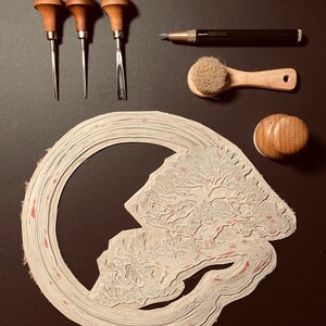 May include: A set of wood carving tools, a brush, a pencil, and a rubber stamp with a detailed design of a tree and a river. The tools include three carving tools with wooden handles and metal blades, a brush with a wooden handle and natural bristles, a pencil with a black barrel and a silver tip, and a rubber stamp with a wooden handle and a detailed design of a tree and a river.