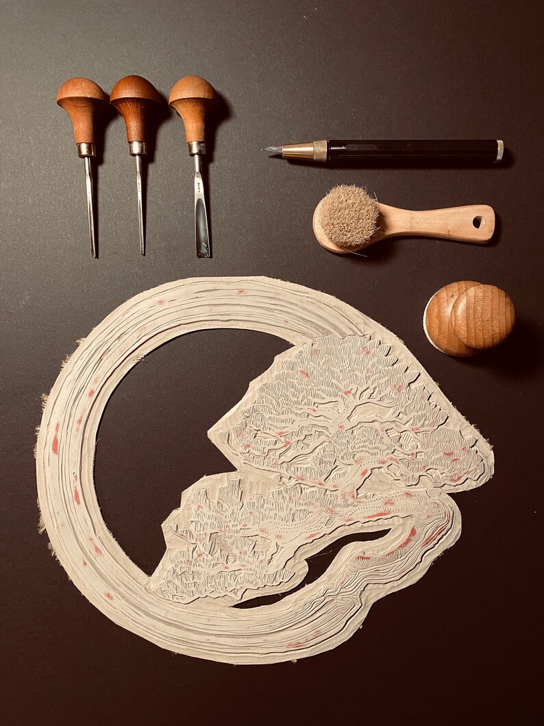 May include: A set of wood carving tools, a brush, a pencil, and a rubber stamp with a detailed design of a tree and a river. The tools include three carving tools with wooden handles and metal blades, a brush with a wooden handle and natural bristles, a pencil with a black barrel and a silver tip, and a rubber stamp with a wooden handle and a detailed design of a tree and a river.