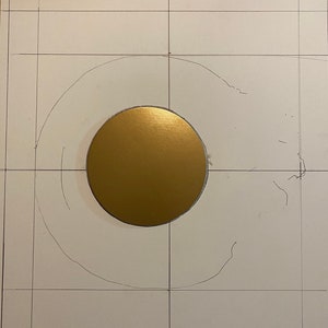 May include: A gold circle is centred on a white surface with a grid pattern. The circle is surrounded by a faint pencil outline of a larger circle.