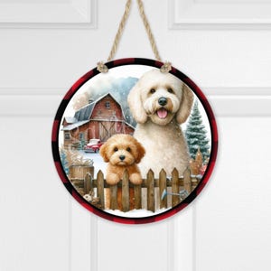 Könnte beinhalten: Runder Holzschild mit einer Aquarellillustration von zwei Hunden in einer Winterszene. Das Schild hat einen rot-schwarzen Büffel-Plaid-Rand und ein Juteseil zum Aufhängen. Eine Scheune und Weihnachtsbäume befinden sich im Hintergrund.
