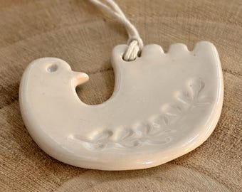 May include: A cream ceramic bird ornament with a textured surface and a floral design. The bird-shaped decoration is suspended by a cream string. The ornament is on a wooden surface.
