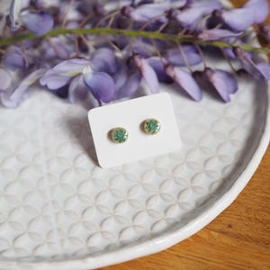 Op de afbeelding: Een paar kleine, ronde oorbellen met een witte achtergrond en groene bloemmotieven. De oorbellen worden gepresenteerd op een wit bord met een decoratief patroon.