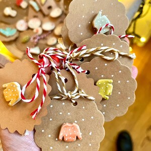 Könnte beinhalten: Ein Satz brauner Papier-Geschenk-Etiketten mit weißen Punkten und kleinen Keramik-Anhängern in Form von Pullovern. Die Pullover sind mit weißen Punkten verziert und in verschiedenen Farben erhältlich, darunter Orange, Gelb und Grün.